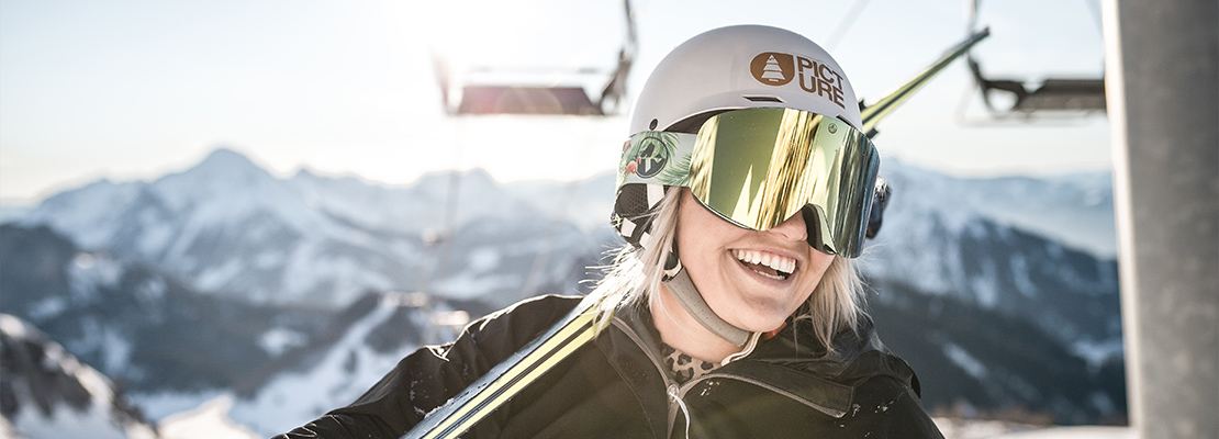 Eine Skifahrerin mit Helm, die Ihre Ski auf den Schultern trägt. Im Hintergrund ein Sessellift und Bergpanorama