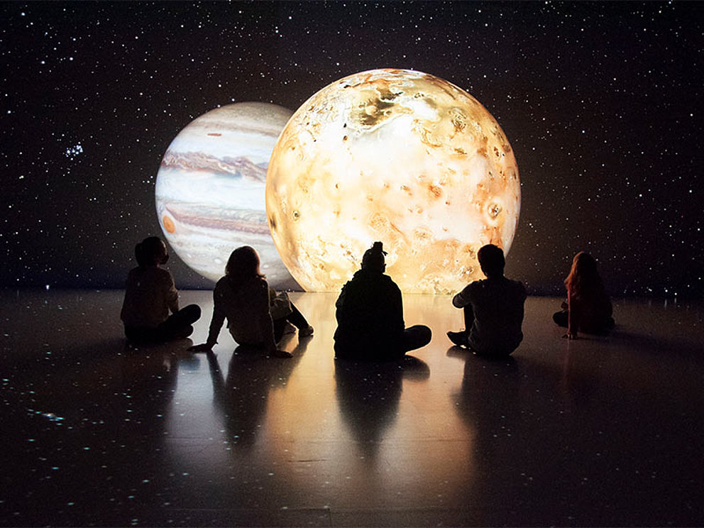 5 Personen sitzen am Boden, im Hintergrund das Weltall auf einer Leinwand