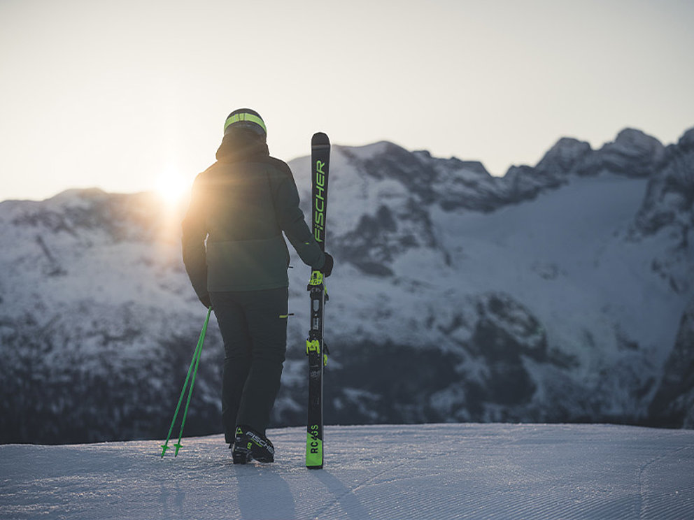  Ein Skifahrer von Hinten, der einen Ski in der rechten Hand hält. Im Hintergrund Bergkulisse.