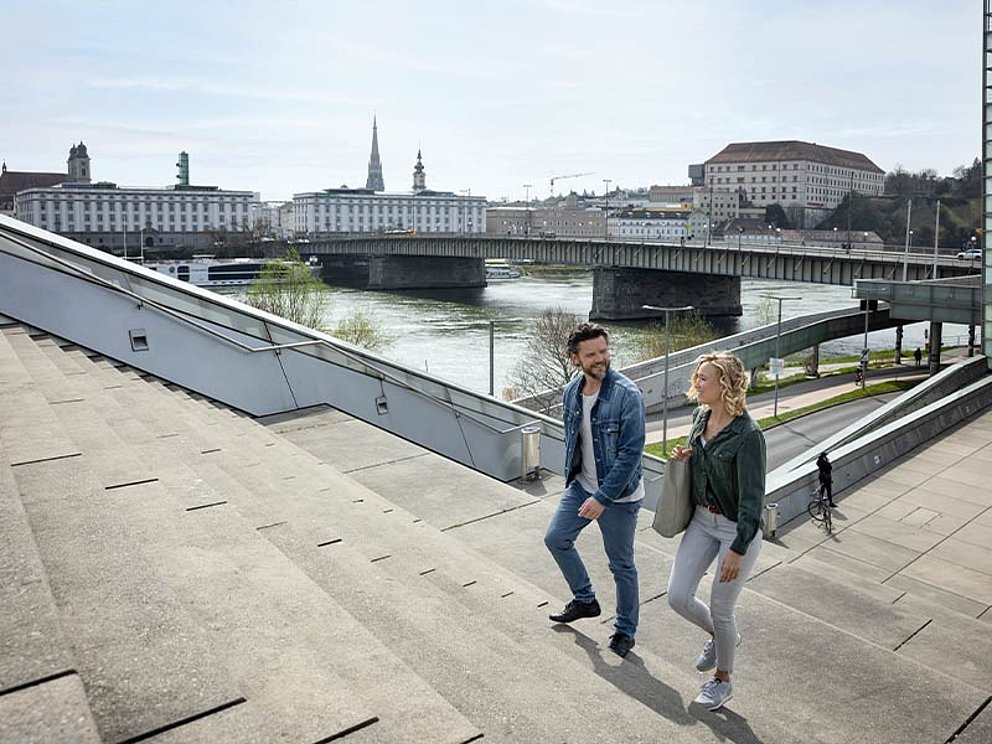 Ein Paar geht über die Treppe beim AEC, im Hintergrund die Donau und die Stadt Linz