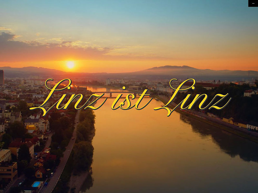 Geschwungener Schriftzug "Linz ist Linz" auf einer Stadtansicht im Sonnenuntergang