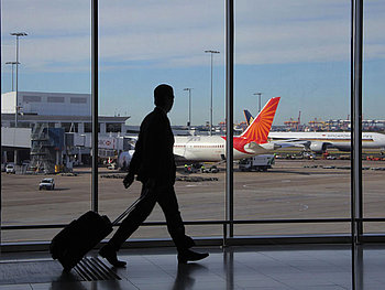 Person mit Rollkoffer vor einem großen Glasfenster, dahinter Rollfeld, Flughafen und Flugzeug