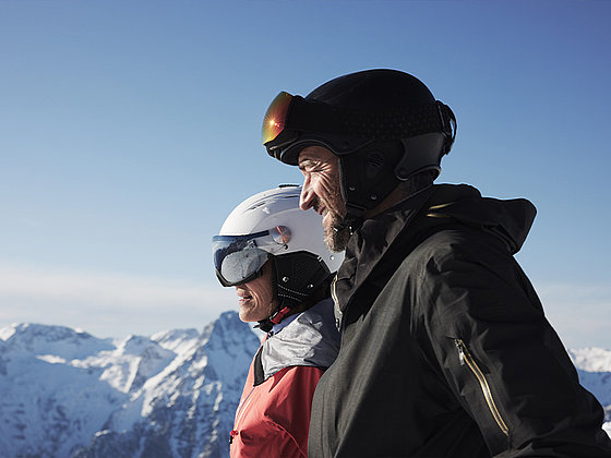 ein Paar mit Skihelm, im Hintergrund verschneites Bergpanorama