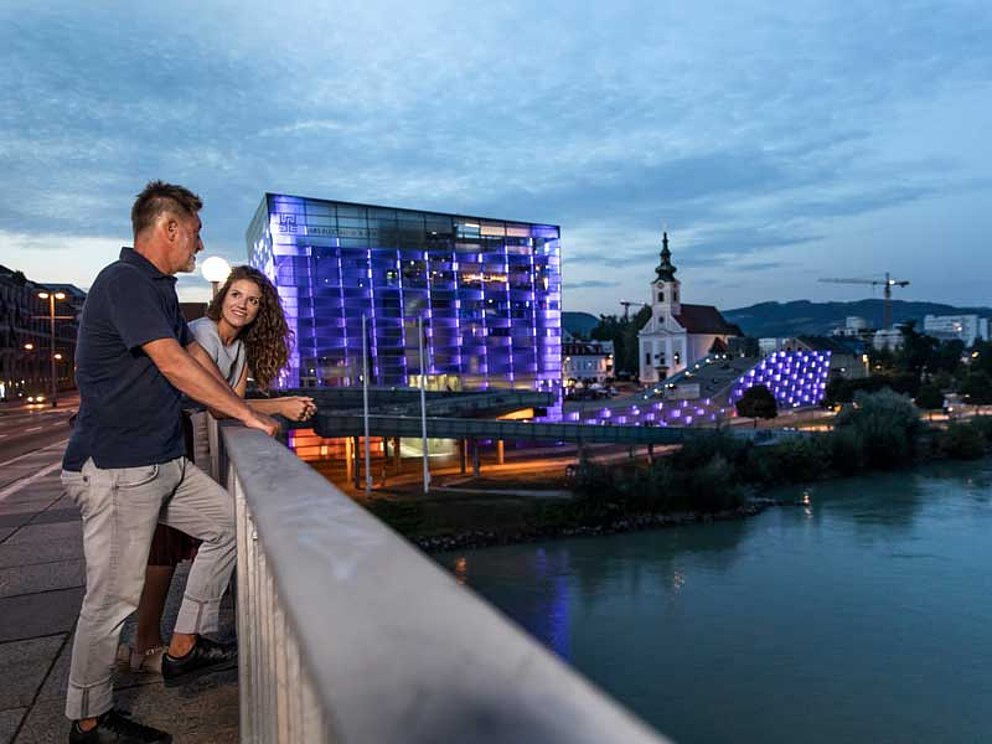Ein Paar steht in der Abenddämmerung auf der Brücke in Linz, im Hintergrund das beleuchtete Ars Electronica Center