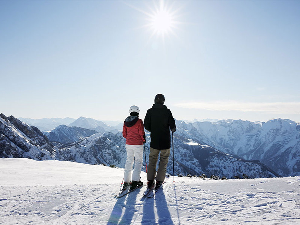 ein Paar in Skiausrüstung von hinten, im Hintergrund verschneites Bergpanorama und Sonnenschein