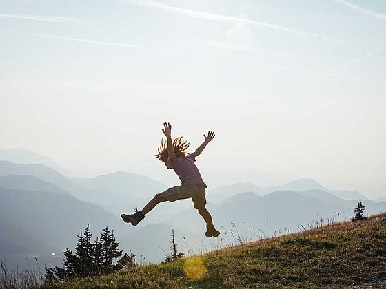 Kind macht einen Luftsprung auf einer Almwiese. Im Hintergrund die Berge.