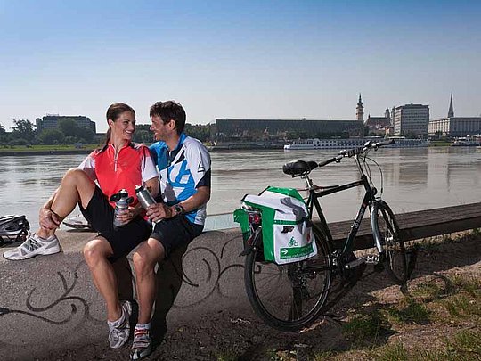 Ein junges Paar sitzt auf einer Mauer am Donauufer in Linz. Rechts von ihnen steht ein Fahrrad. Am anderen Ufer grüßt die Silhouette der Stadt.