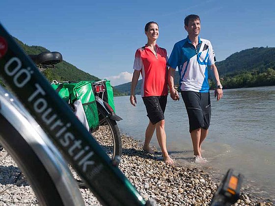 Ein junges Paar watet barfuß in Raddressen im seichten Donauufer. Davor stehen ihre Fahrräder.