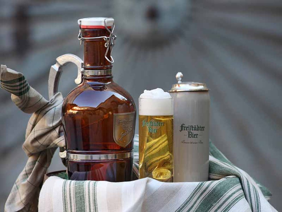 Auf einem stehenden Fass befinden sich ein schaumgekröntes Glas Bier, ein Humpen und eine bauchige Bierflasche.