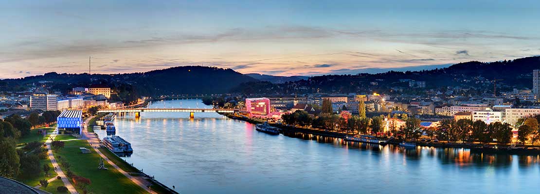 Blick auf die Stadt Linz mit Donau bei Dämmerung. AEC und LENTOS Kunstmuseum leuchten bunt zu beiden Seiten der Donau.