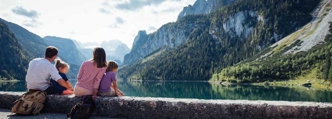 Eltern und 2 Kinder sitzen auf einem Mäuerchen und blicken auf einen Gebirgssee, umrahmt von hohen Bergen