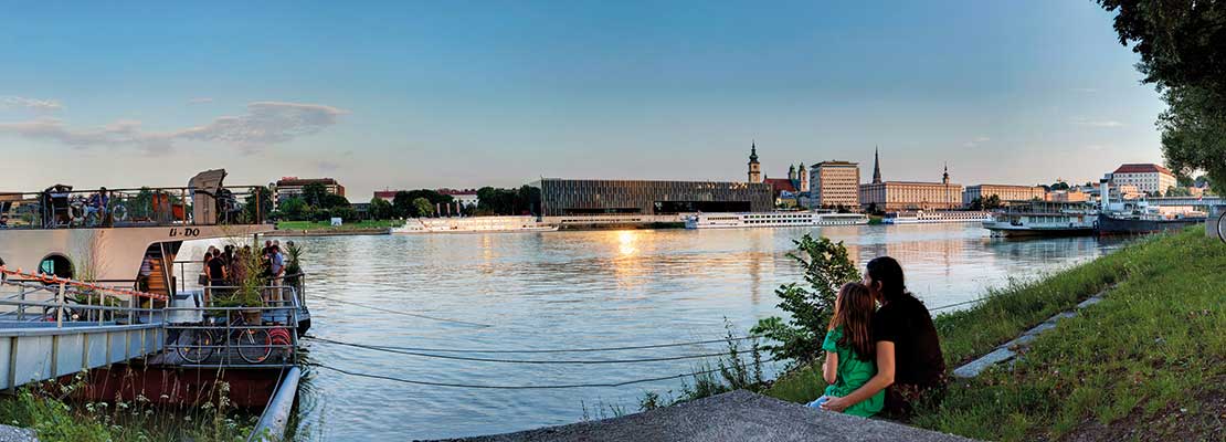 Paar sitzt am Donauufer in der Wiese. Daneben ein Lidodeck mit fröhlichen Menschen. Am anderen Ufer Linz.