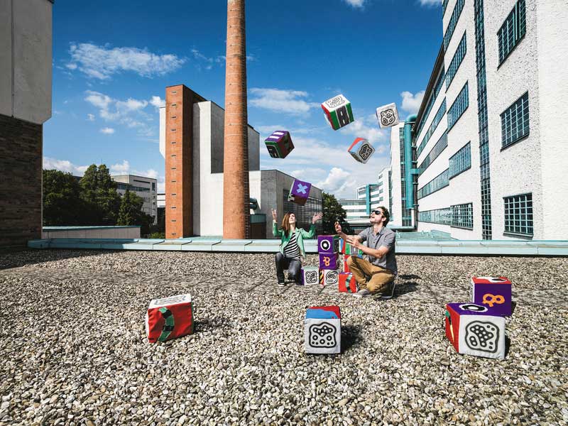 2 Menschen jonglieren mit Würfel auf einem Flachdach in industriellem Umfeld